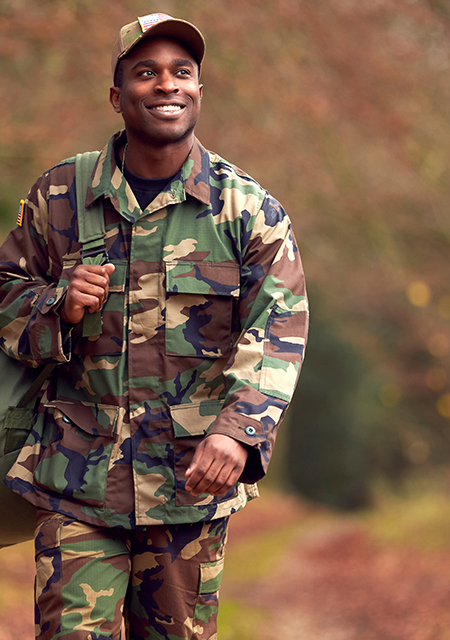 A smiling army officer with a backpack on his shoulders, walking outside.
