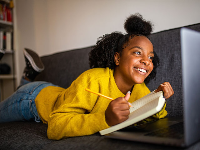 Smiling African American young woman laying on the couch on her stomach, holding a pencil and notebook, taking notes from her laptop in front of her.