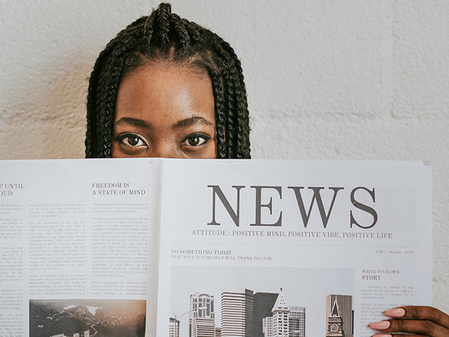 A young black woman reading/ holding a newspaper. The newspaper covers the lower-half of her face. Only her eyes are visible
