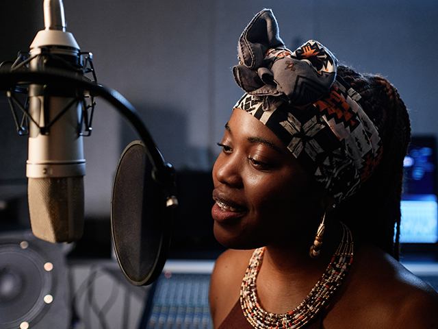 Smiling young African American female singer standing in front of microphone recording song in studio