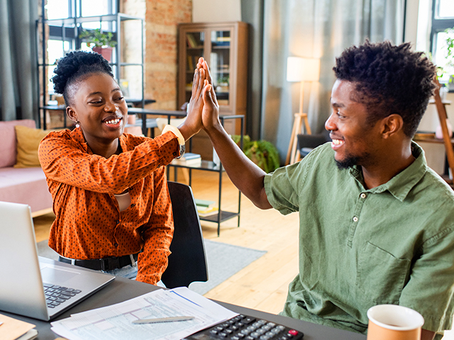 Happy young African American male and female college classmates colleagues giving each other high-five while working together at table