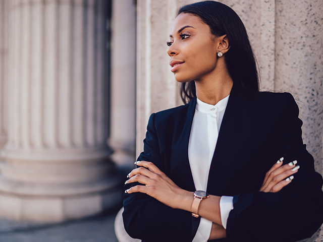 Confident looking Female African American woman lawyer dressed in elegant black suit folding arms and looking to the side, standing in front of white government building.
