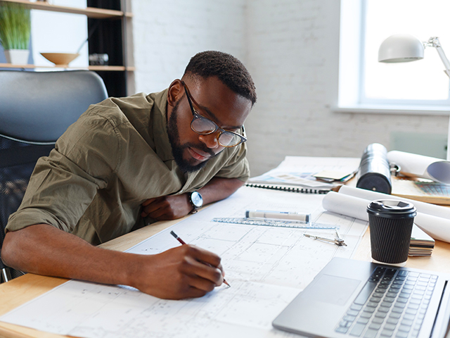 African American male architect working in office with blueprints, sketching an architectural plan.