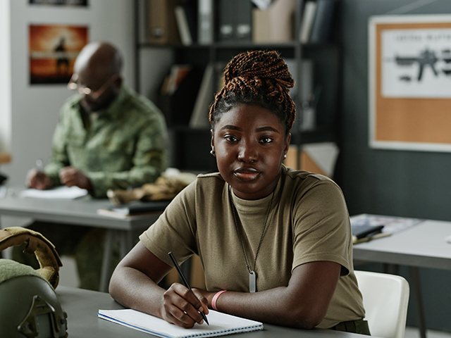 African American female student in casualwear sitting by desk with military equipment and looking at camera while making lecture notes